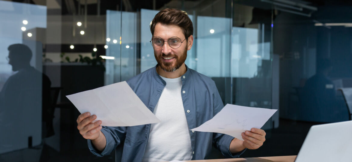Satisfied with the result financier businessman holds in his hands and examines contracts and reports smiling and happy, man in a casual shirt works inside the office using a laptop in paperwork